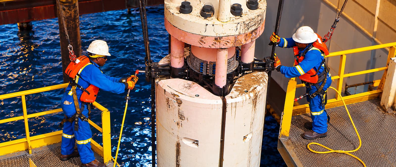 Two crew members in harnesses and hard hats working at a wellhead on an offshore platform.